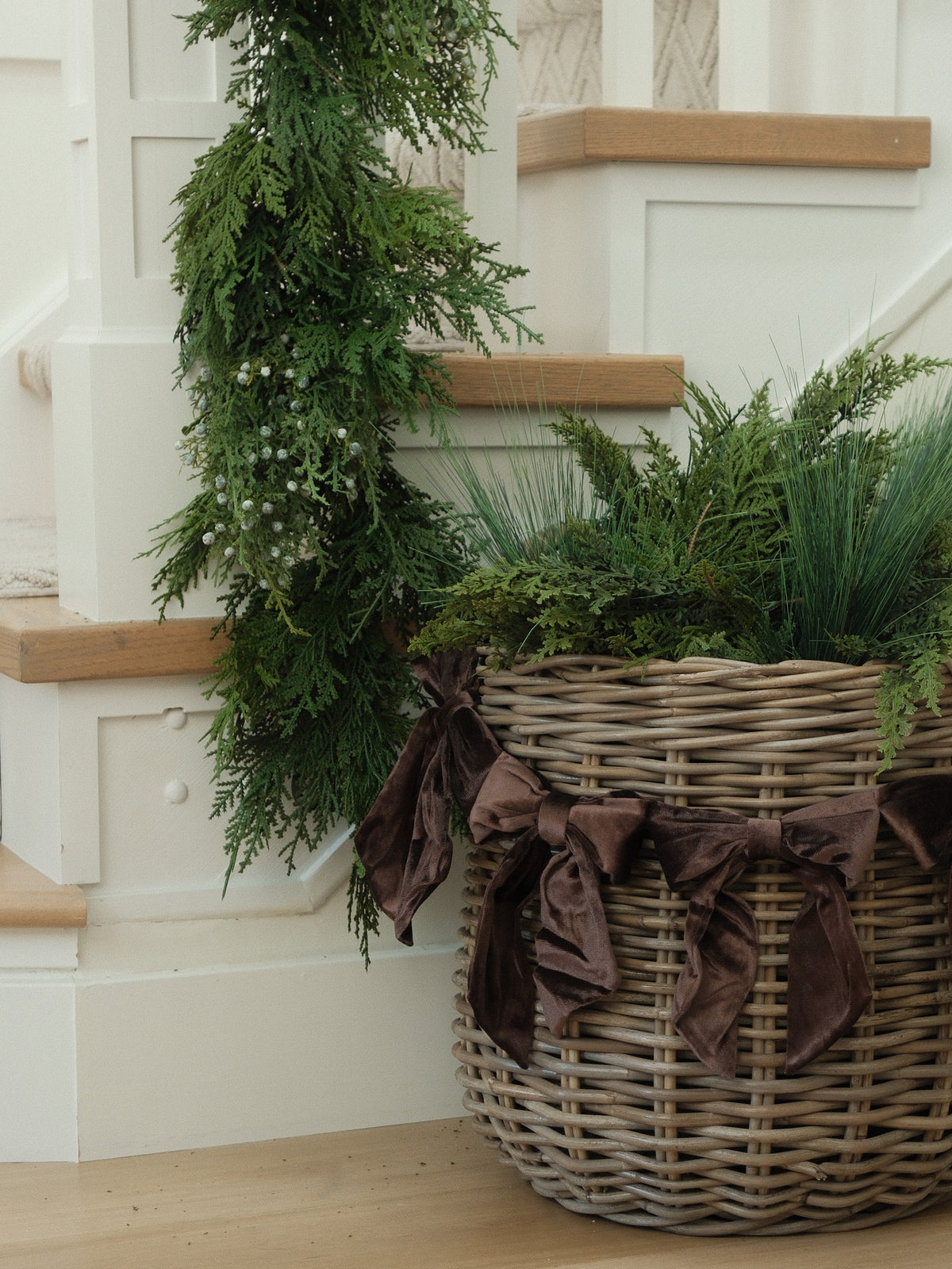 A large woven wicker basket filled with green holiday foliage and garlands, tied with dark brown velvet bows, placed on a wooden floor beside white stairs.